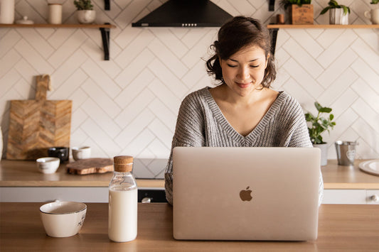 A woman sits at a kitchen counter working on a laptop and smiling