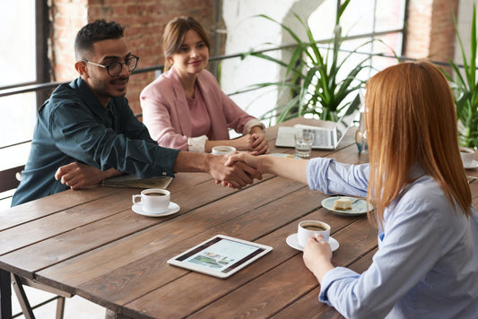 A man and woman sit across a wooden table from another women, who is shaking hands with the man