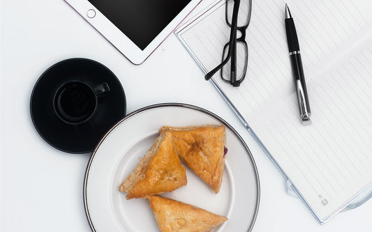 A collection of morning things: a cup of coffee, small pastries, a notebook, pen, reading glasses, and an iPad.