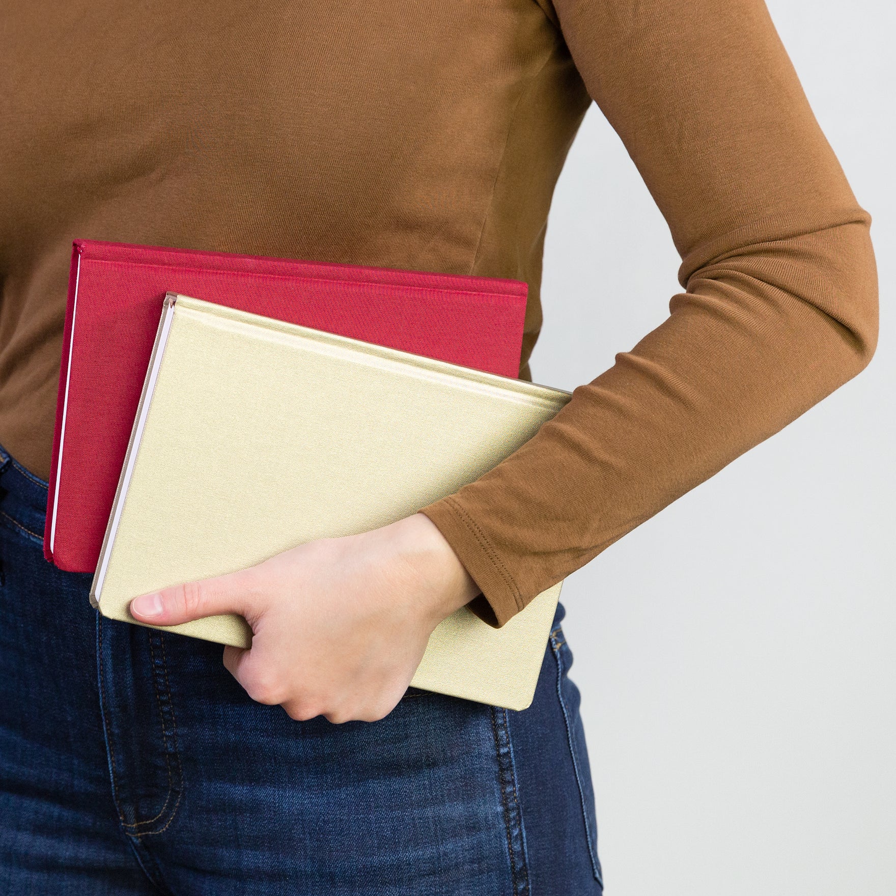 Side view of a woman in a brown shirt and blue jeans holding two red and gold planners against her hip.