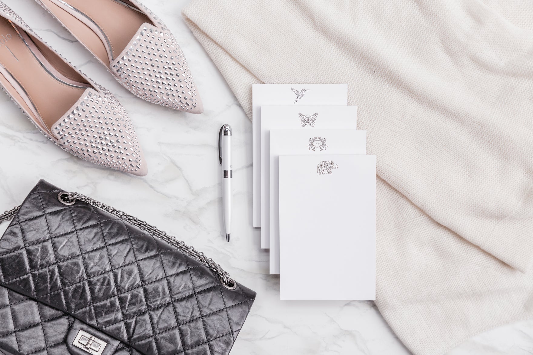 A gold foil notepad sits on a marble surface next to a quilted black handbag, fountain pen, and flats.