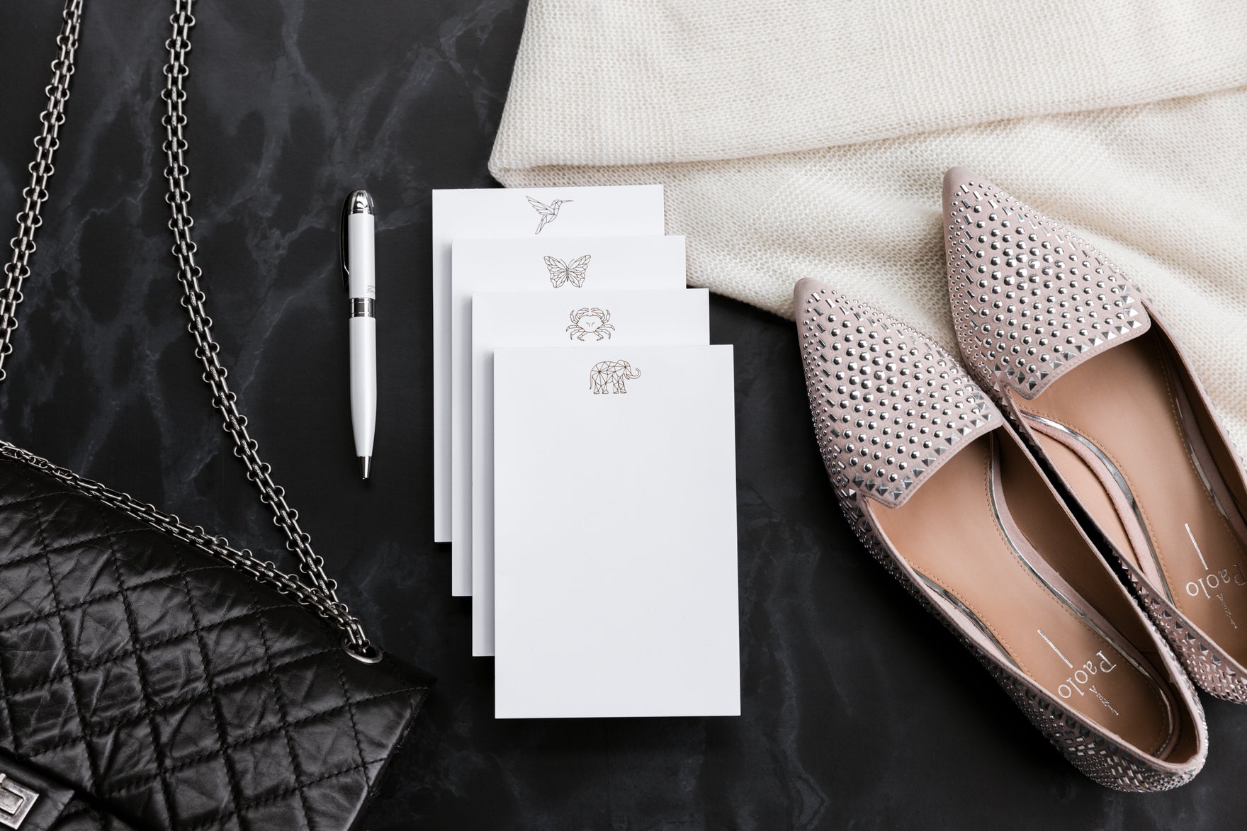 A quilted black handbag, a pen, gold-foil print notepads, and a pair of elegant flats sit on a black table.