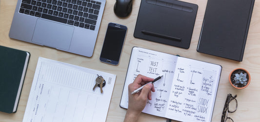 An array of desk supplies including a filled out planner with a hand holding a pen