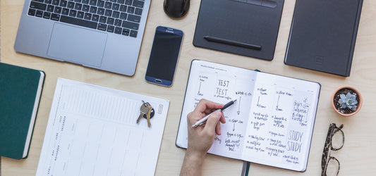 A student planner open on a full desk with keys, glasses, computer, and phone.