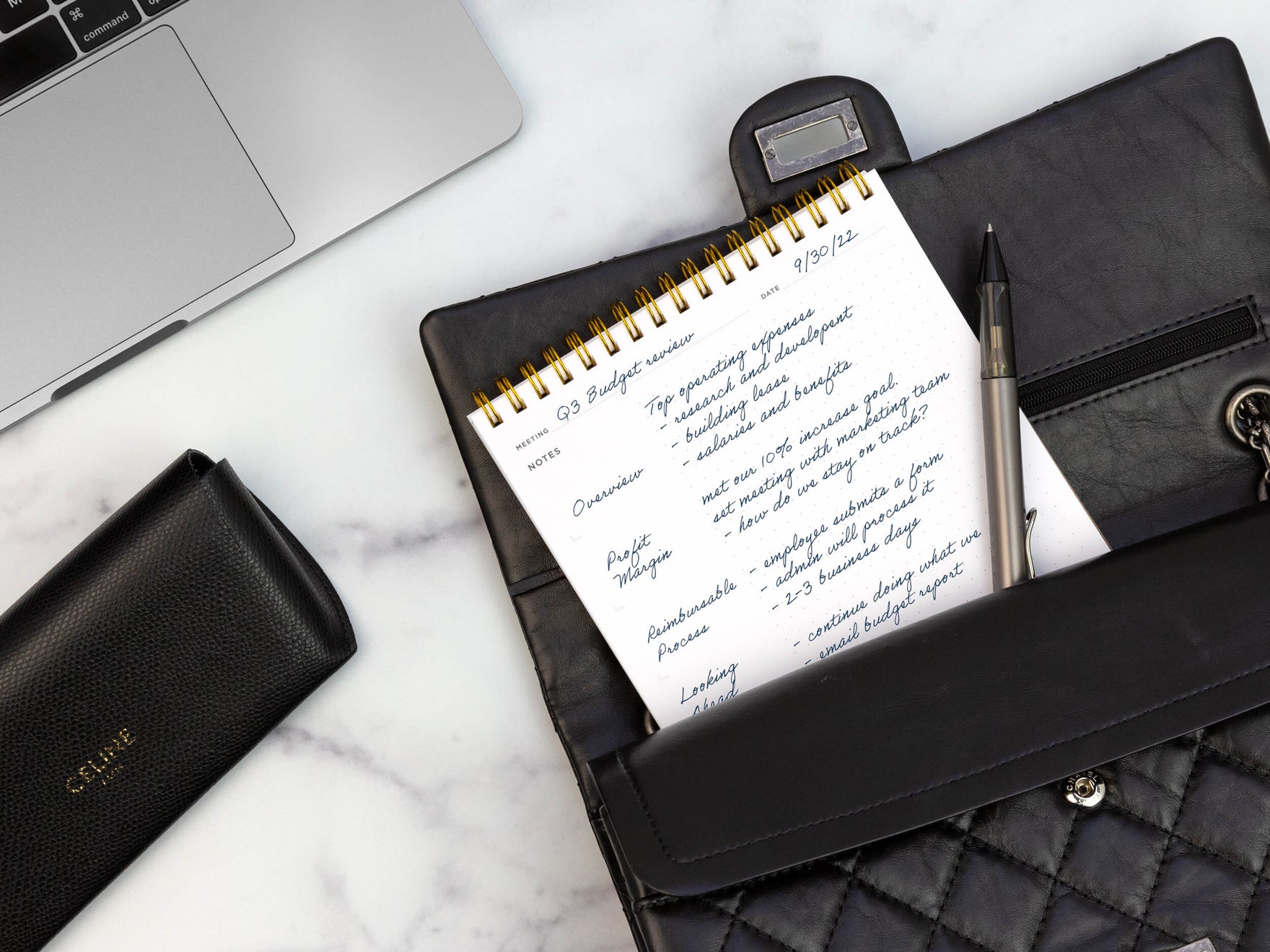 A meeting notes pad sticks out of a black handbag with a silver pen on a white marble table