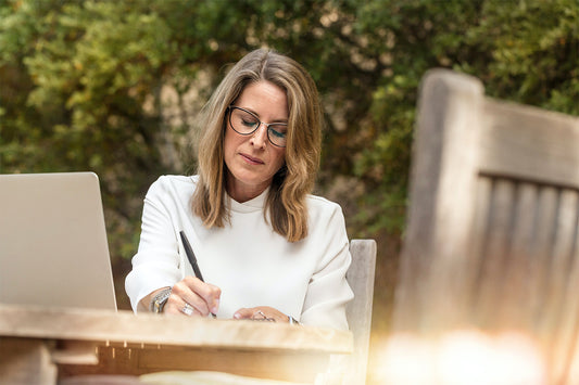 A woman with glasses sits at an outdoor table and writes in a notebook