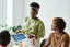 A woman stands and shows a document to two seated people in an office
