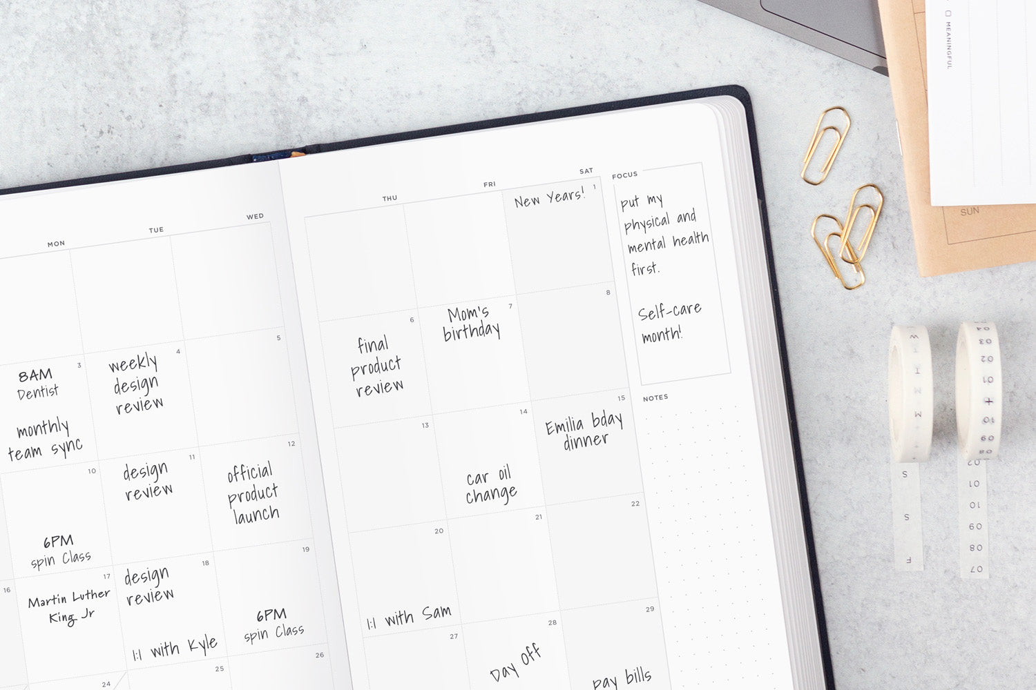 A planner with many appointments written in it, sitting on a white table with gold paperclips and other accessories