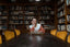 A woman at a long dark table with gold velvet chairs looks down at the table, with filled bookshelves behind her