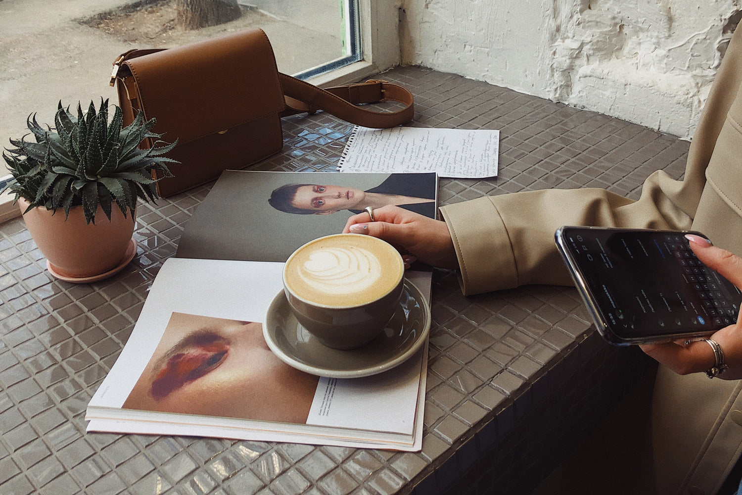 A latte in a mug sits on a magazine on a table with a plant and a handbag in front of a window