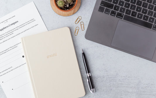 A gratitude journal and a gratitude worksheet sit on a desk next to a pen, a plant, and a computer.