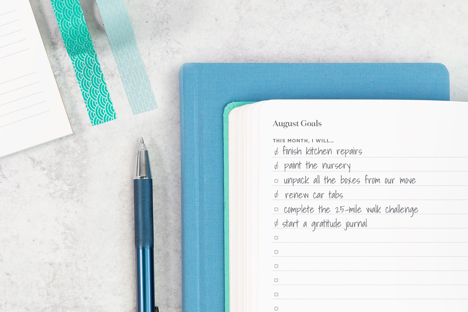 A blue planner on a white table next to a blue pen and teal washi tape. In the open planner is a list of goals.