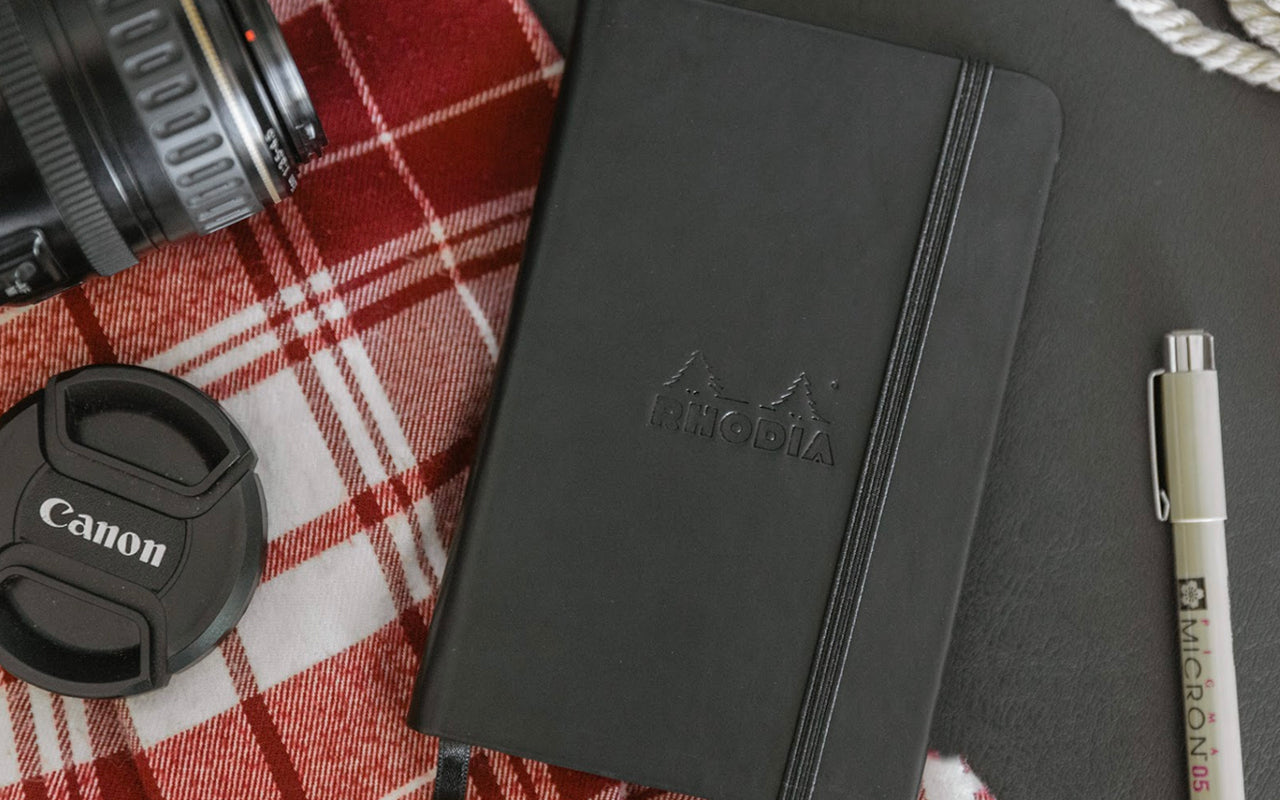 A Rhodia notebook sits on a plaid blanket with a camera lens and lens cover