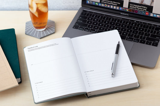 A planner sits open on a desk on top of a laptop and next to a glass of iced tea.