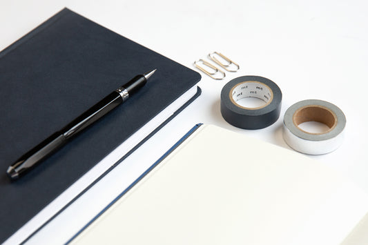 A weekly deskpad sits on a white desk next to a cup of coffee, a phone, gold foil notepads, and some greenery.