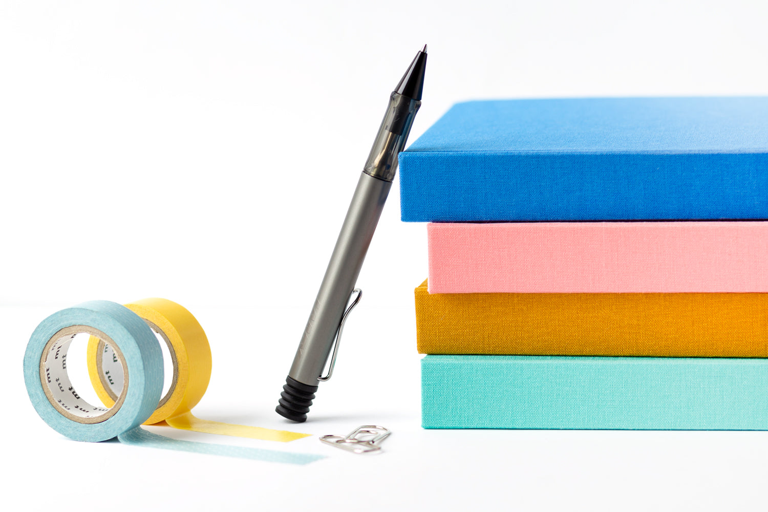 A stack of colorful planners sit on a white background with a pen and two rolls of colorful washi tape.