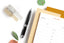 An array of cute office supplies for fall, including a pen, a decision notepad, grey and silver washi tape, and a goldenrod ink+volt planner.