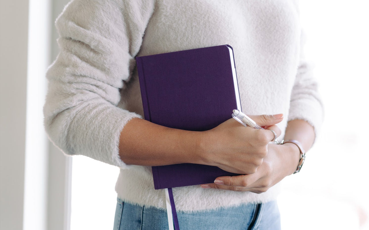 A person holds a purple planner and a pen, standing in a determined way.