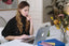 A woman sits at a laptop on a white desk with flowers