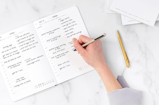 A hand holding a pen fills out a daily to-do list notepad on a white desk.