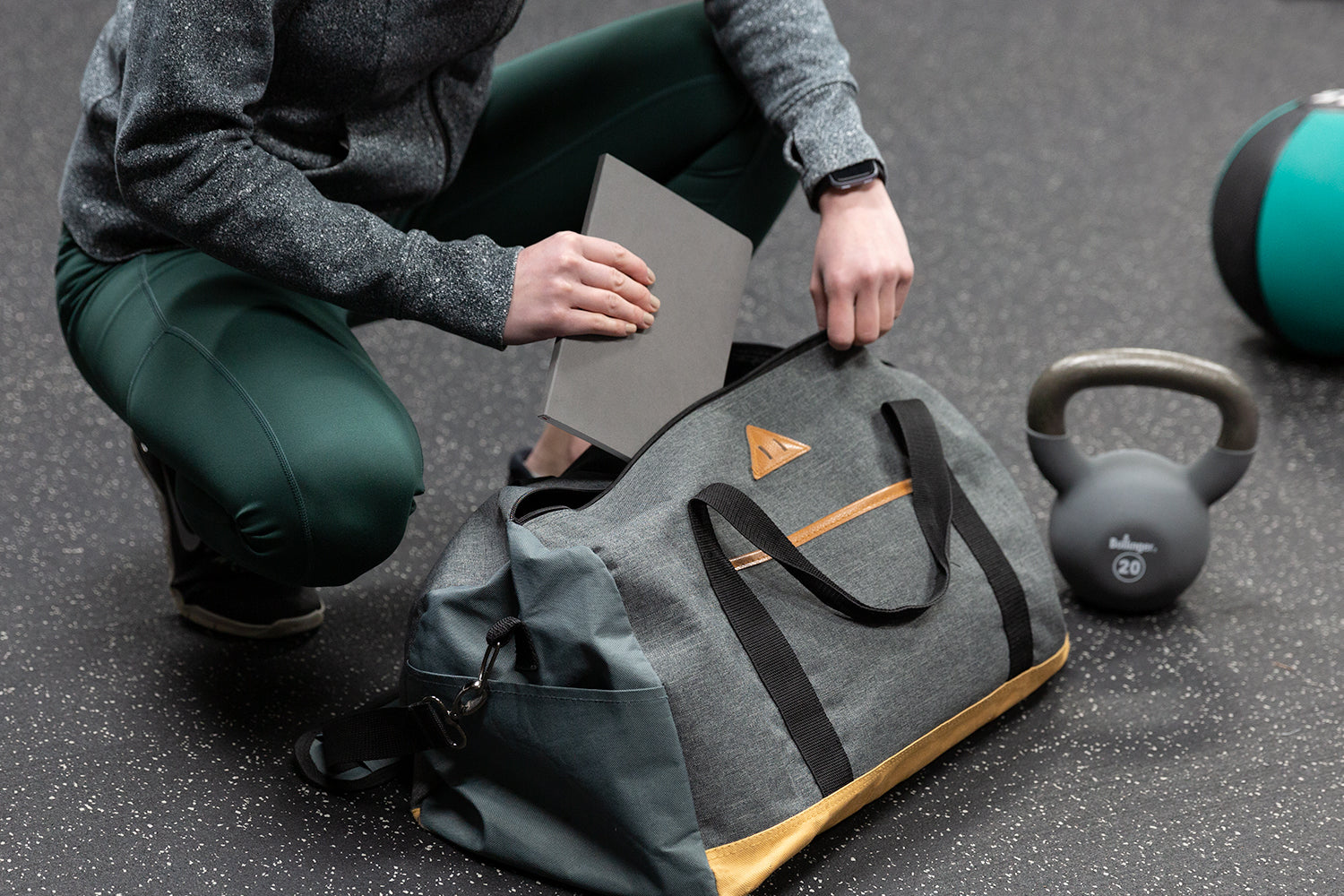 A woman in workout clothes puts a grey fitness journal into a grey gym bag.