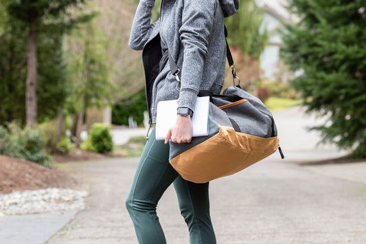 A person in green workout pants and a grey sweater holds a fitness journal next to a grey gym bag over their shoulder