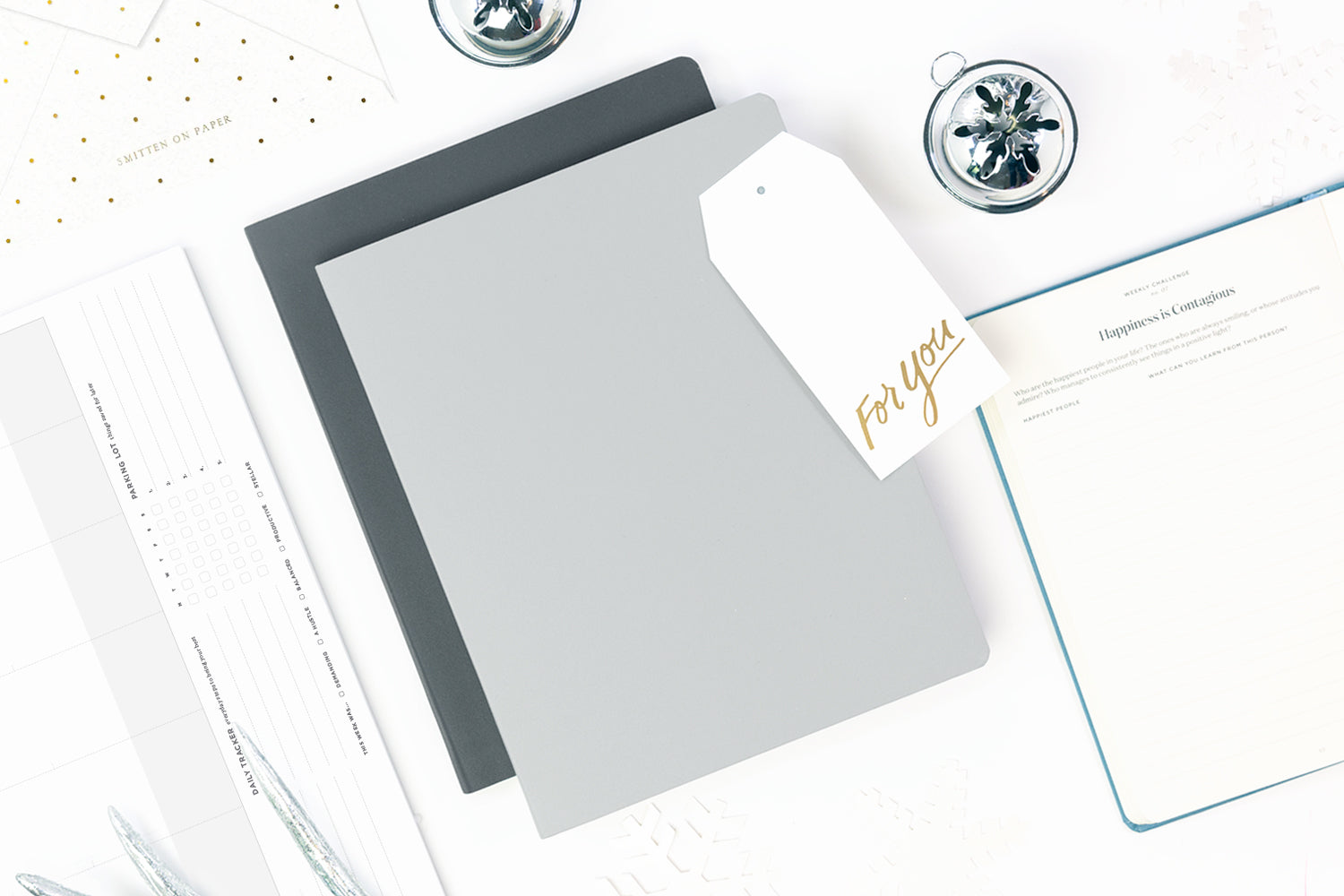 An array of elegant office gifts: a selection of grey notebooks, a white gift tag, and a slate blue journal open on a white tabletop.