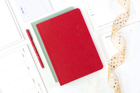 A red planner on top of an array of notepads, next to a red pen and a gold ribbon.