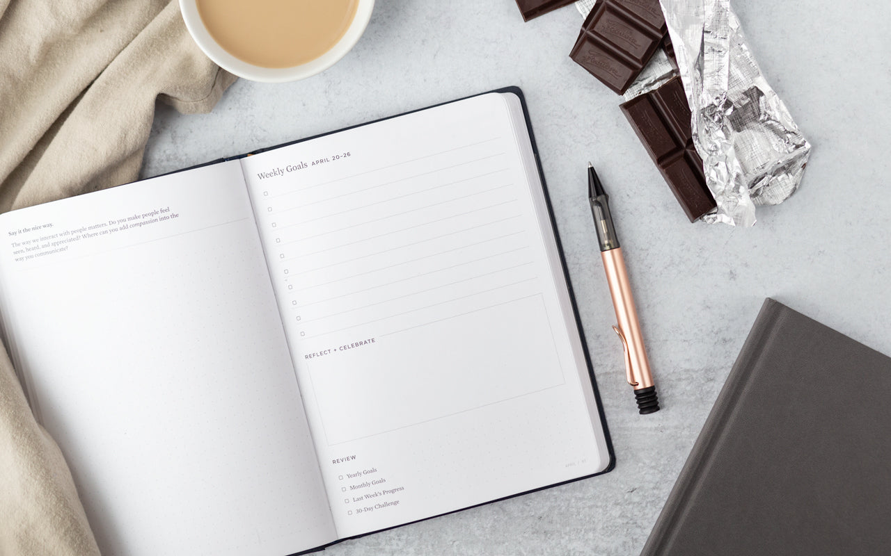 A planner sits next to a rose gold pen and a chocolate bar on a table with a cup of tea and cozy blanket.