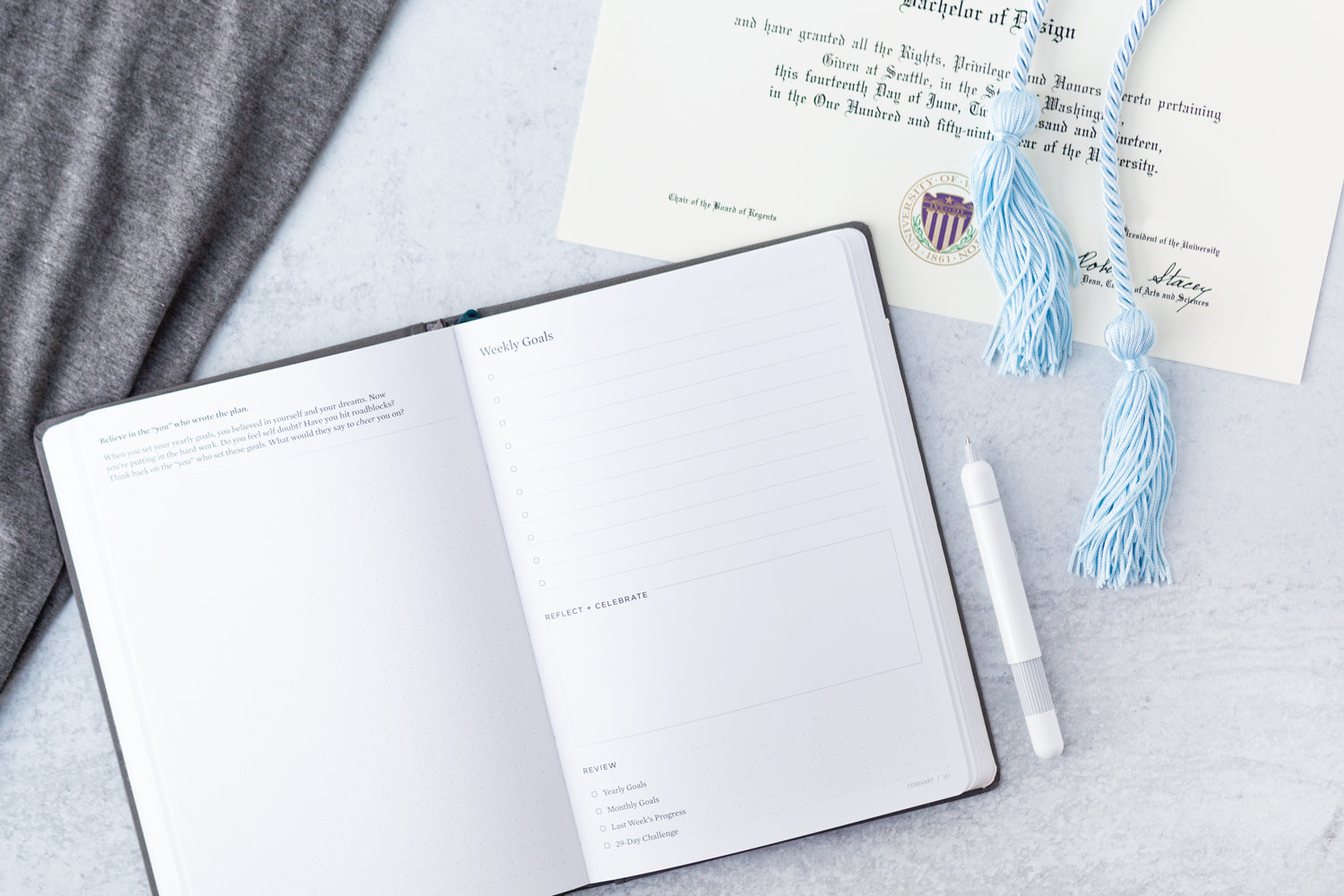 A college graduation cap's blue tassels lie on top of a diploma, next to an Ink+Volt Planner and white pen.