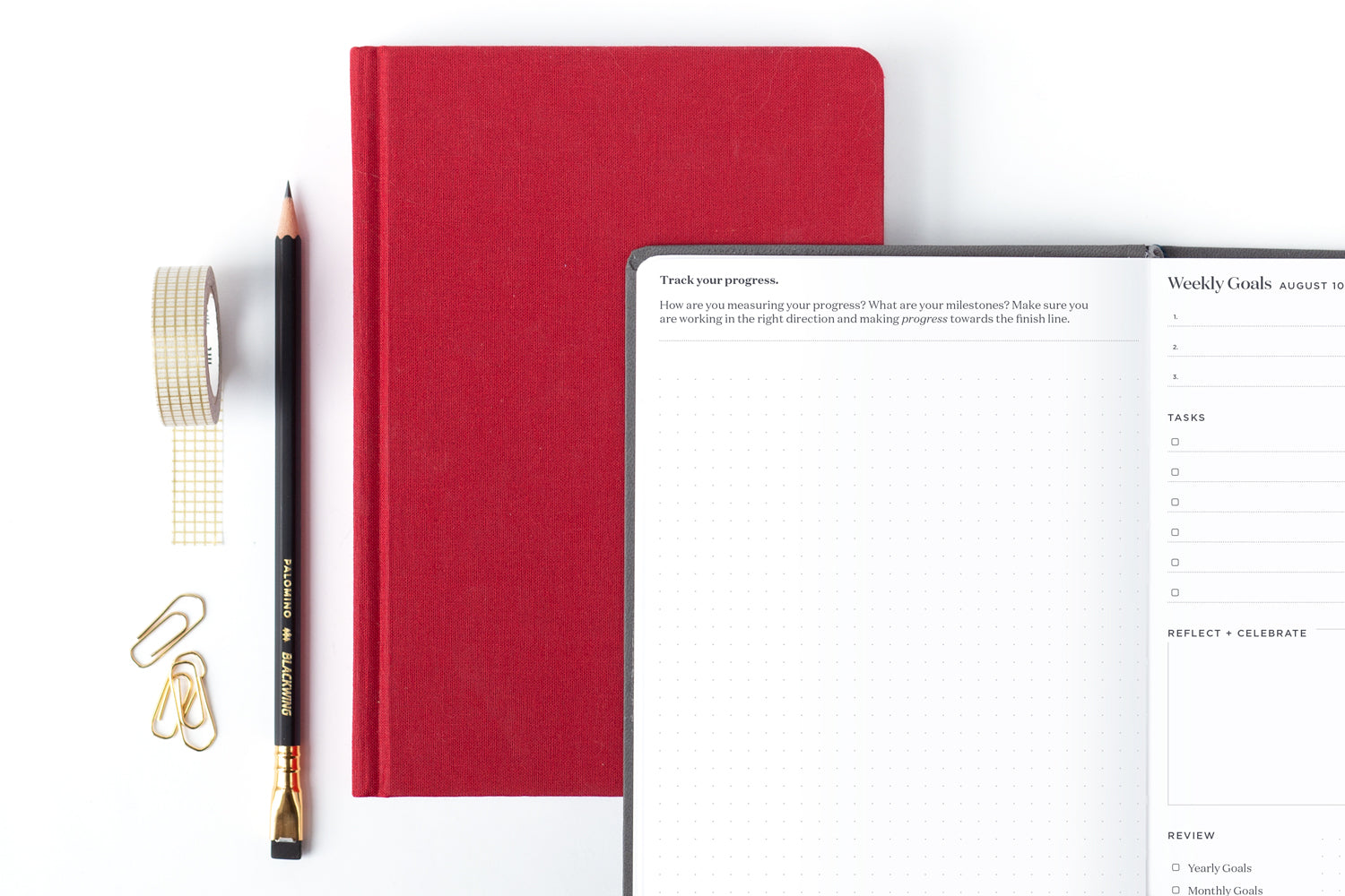 A red planner sits on a desk under another open planner, plus a pencil, washi tape, and gold paper clips.