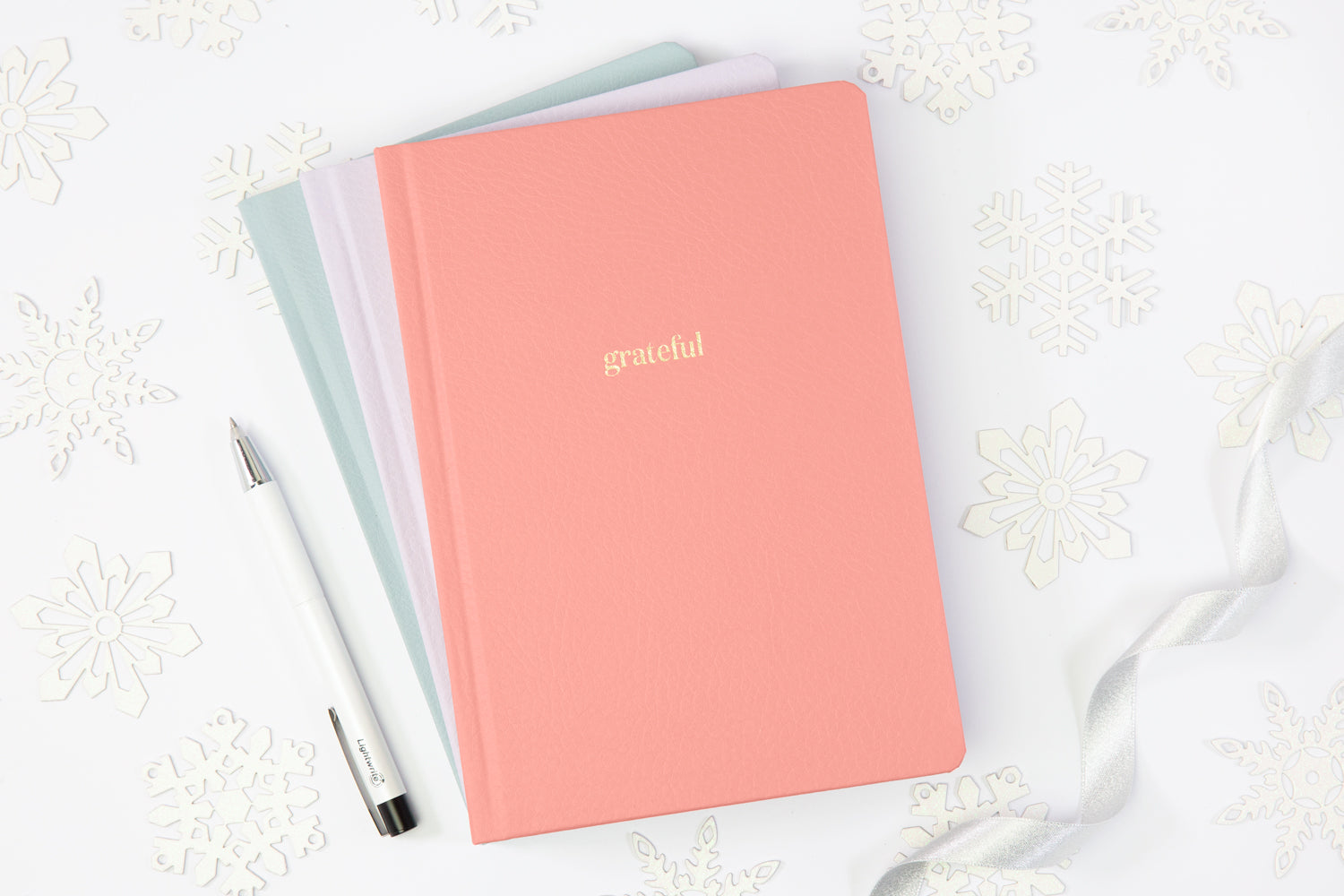 A stack of pink, blue, and lavender gratitude journals on a white table, with a white pen.