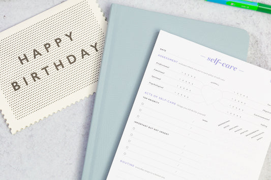 A birthday card, gratitude journal, and self-care notepad sit on a grey table.