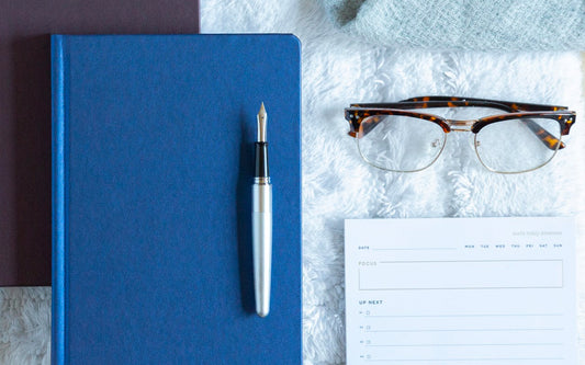 A silver Pilot Metropolitan fountain pen sits on a blue Ink+Volt 2020 Planner Limited Series, next to the Ink+Volt Task Pad and a pair of tortoiseshell eyeglasses