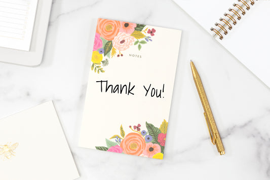 A floral thank you card on a white surface next to a gold pen