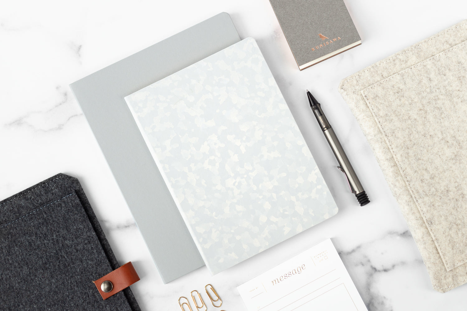 A marble desktop with luxurious office supplies on it including a Founders Notebook, wool planner sleeve, and gold paper clips.
