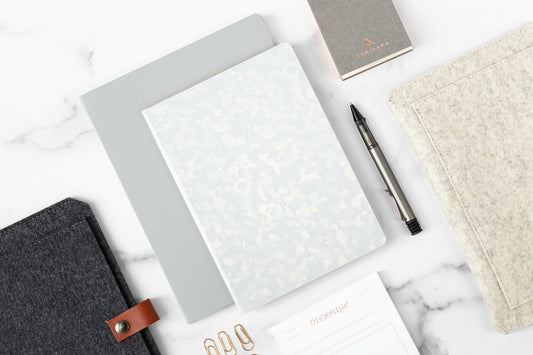 A marble desktop with luxurious office supplies on it including a Founders Notebook, wool planner sleeve, and gold paper clips.