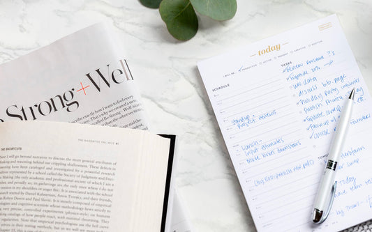 A Today pad is filled out with a to-do list next to a book and a magazine on a marble countertop.