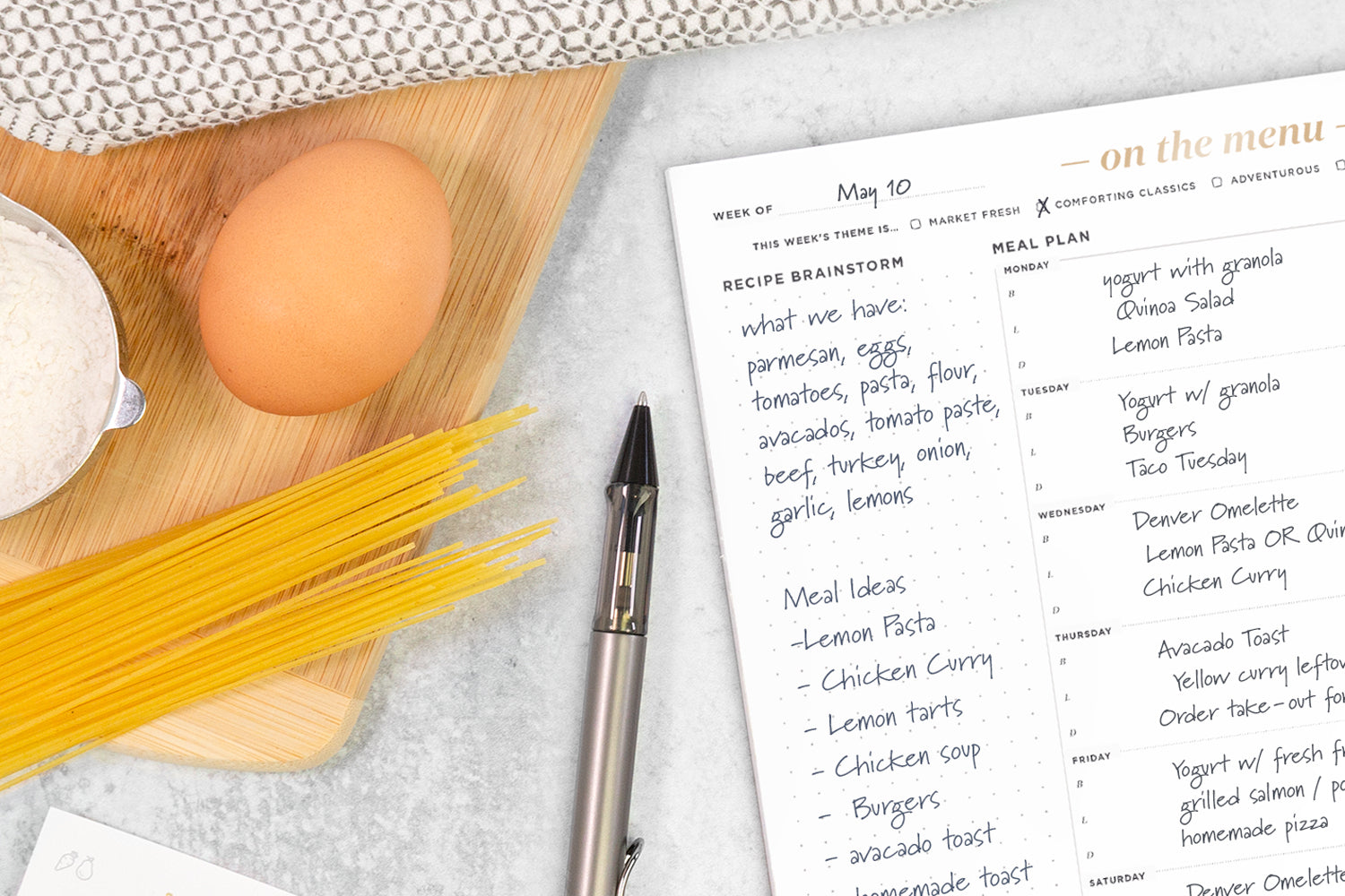 A meal planning pad on a white counter next to a pen, an egg, dried pasta, and a wooden cutting board.