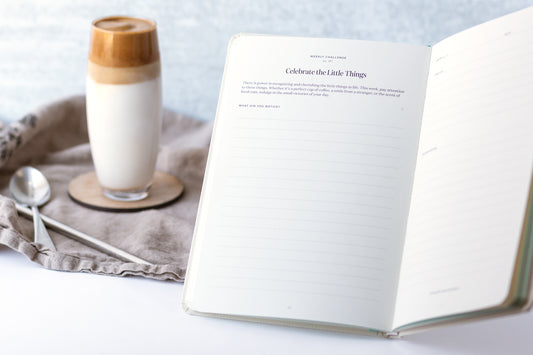 A mindfulness gratitude journal sits open on a table next to a fancy coffee in a tall glass.