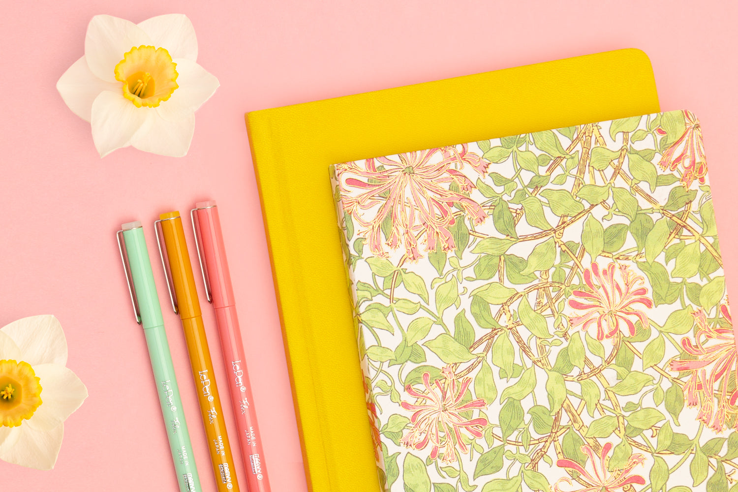A floral notebook sits atop a yellow notebook, next to colorful pens and daisies on a pink table
