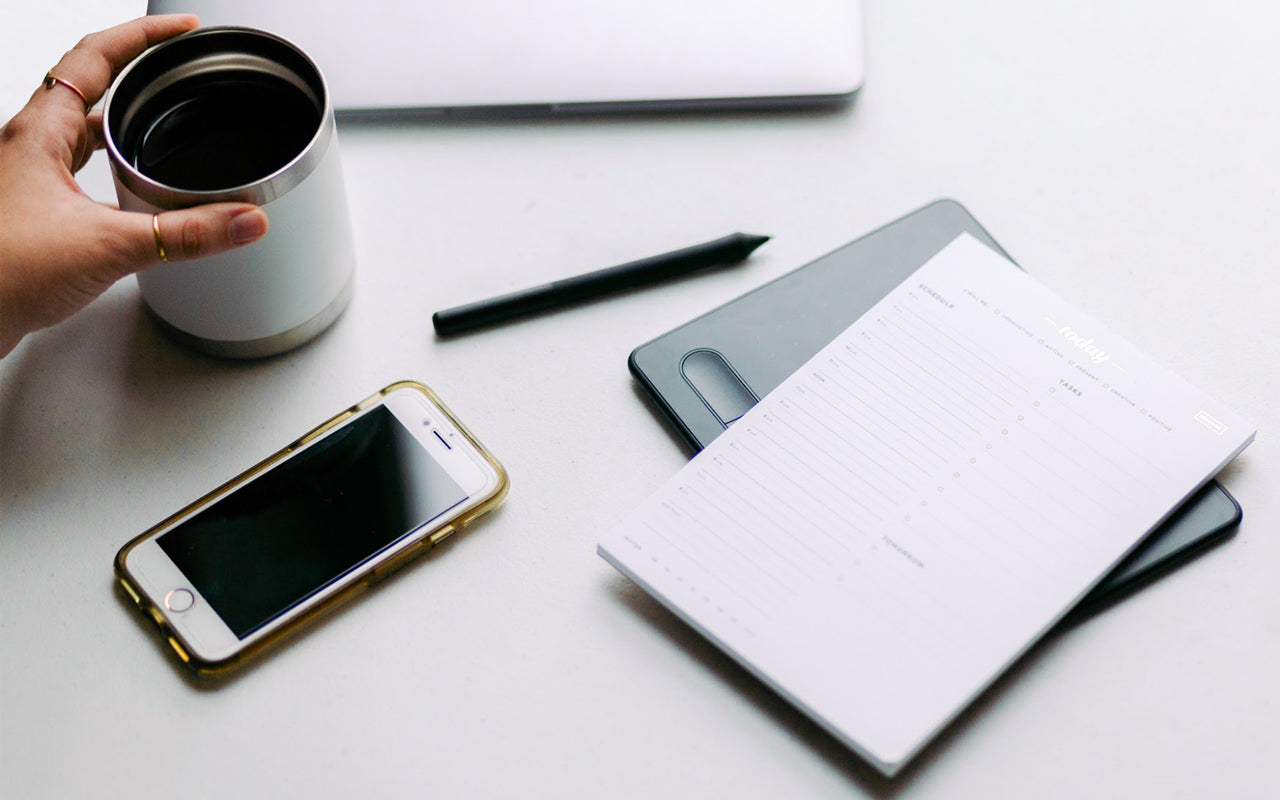 A white desk with a coffee cup, a phone, a notebook, a pen, and a daily organizer notepad.