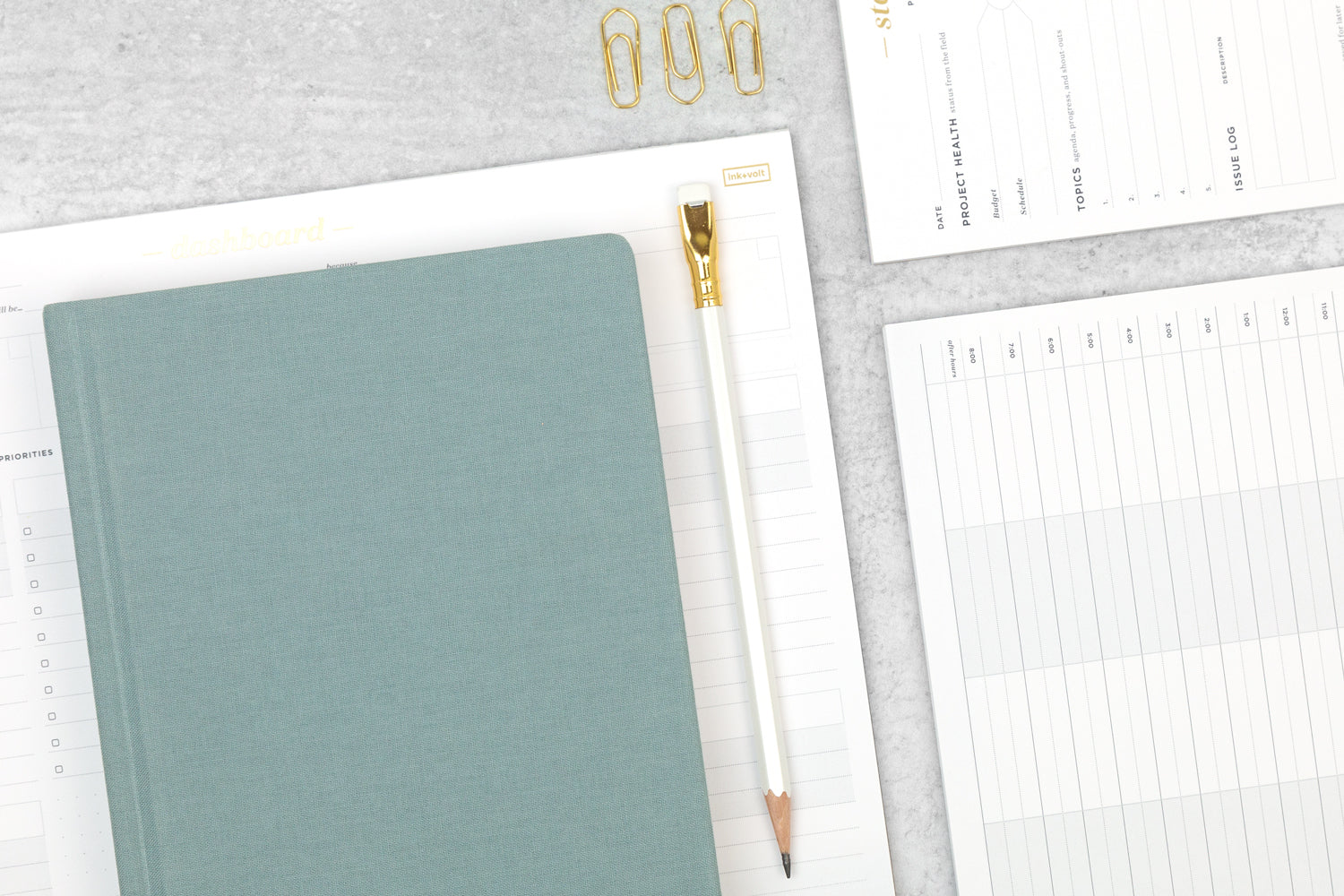 An array of white, gold, light blue planning tools and notepads on a grey desktop.