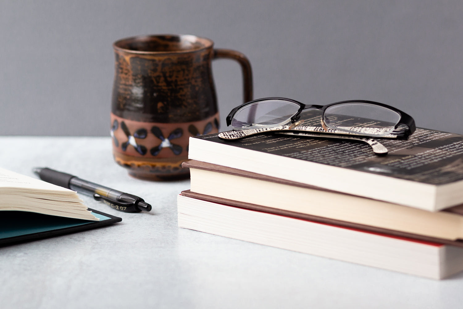 A stack of books sits on a white desk next to a pen and a unique coffee mug.