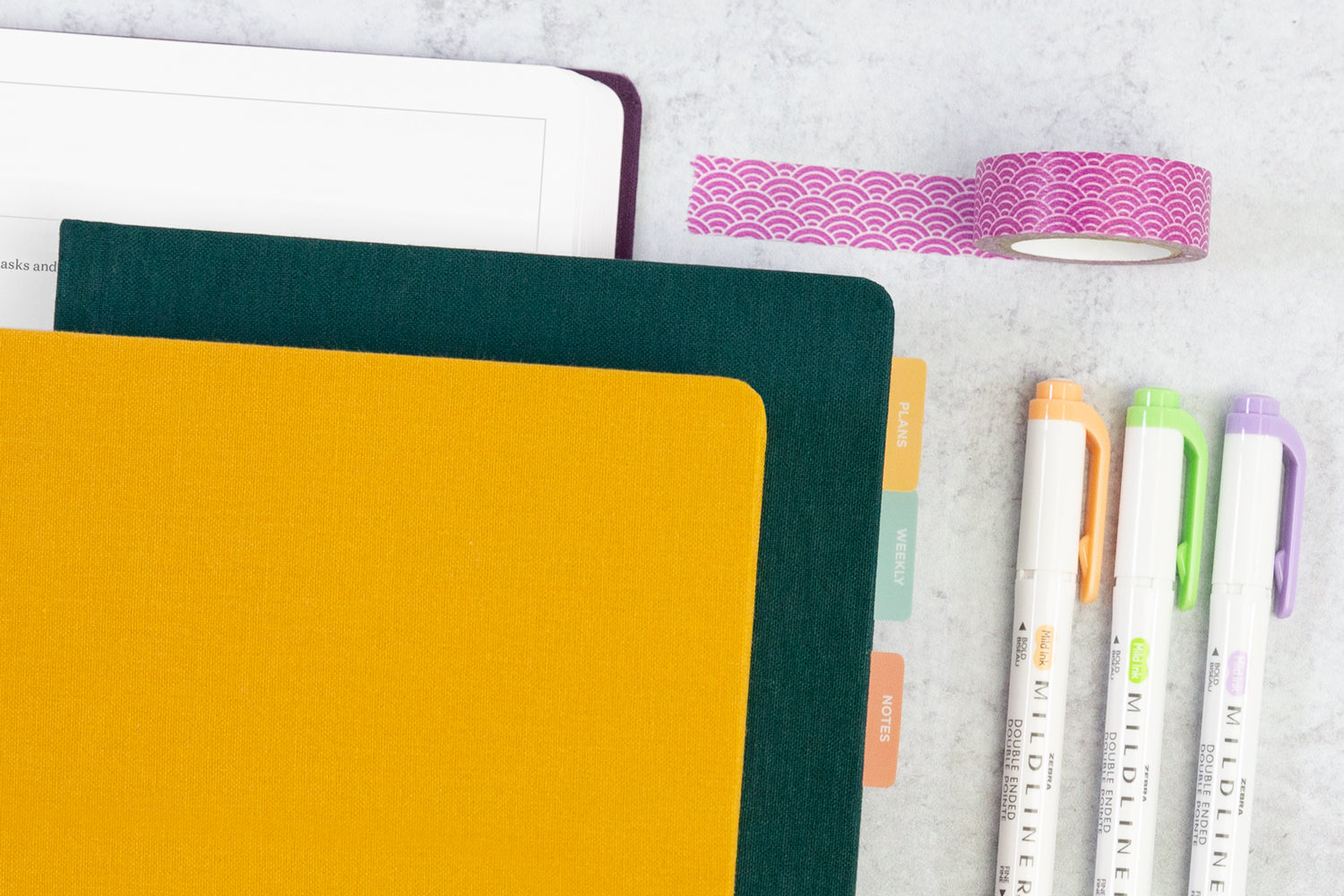 Three planners, stacked, on a white table, next to 3 colorful markers and a strip of pink tape.