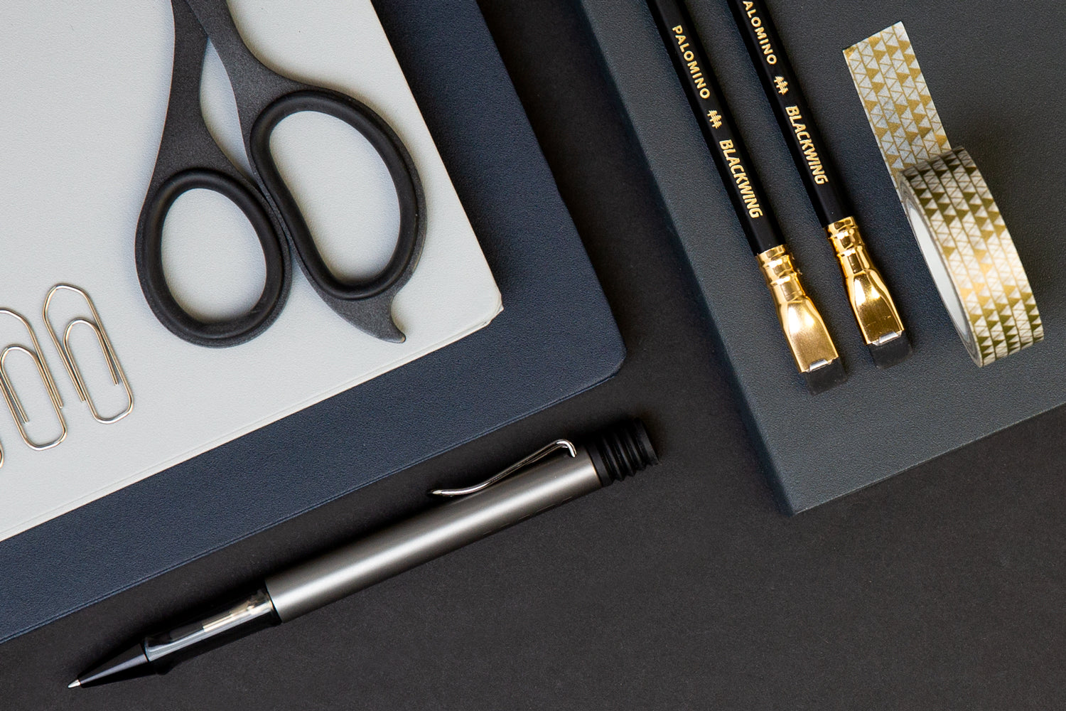 An array of dark colored desk accessories - black scissors, black pencils, a navy planner, and gold washi tape