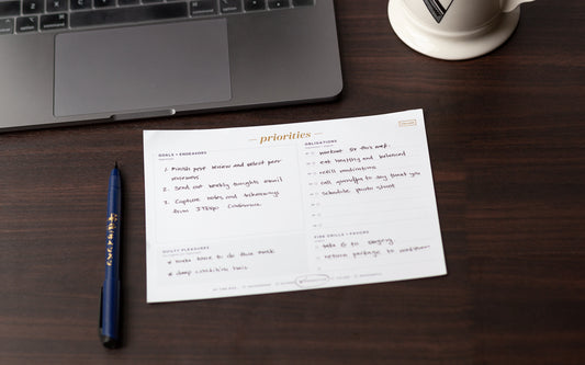 A priority notepad sits on a wood desk next to a blue pen and a laptop.