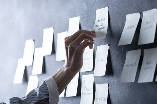 A chalkboard with an array of post-it notes and a hand reaching up to one