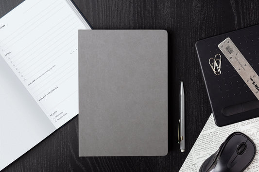 A project planner notebook sits on a desk next to an open planner, paper clips, pen, and other organizational tools.