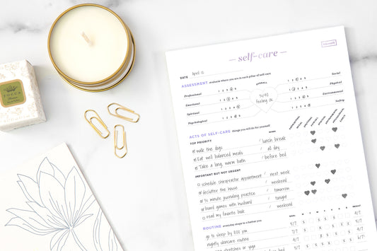 A self-care notepad on a white table next to a candle and gold accessories.